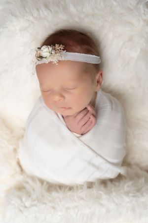 Newborn wrapped in white swaddle wearing delicate floral headband on fuzzy white background.
