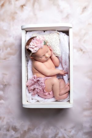 Sleeping newborn with pink accessories photographed in white wooden box from above.