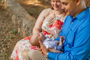 Family sitting together outdoors with newborn baby in blue outfit on blanket with trees in background.