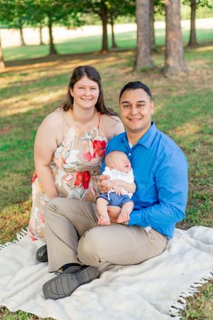 Family of three sits on white blanket for outdoor portrait session in park setting.