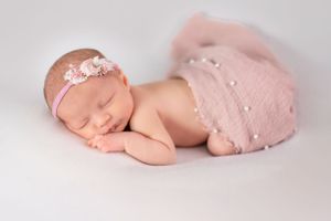 Newborn wearing pink headband and wrap lying peacefully on soft white background during professional photo session.