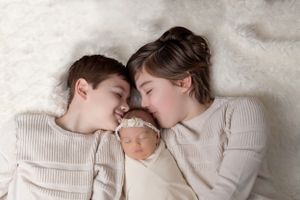 Siblings in matching sweaters sharing sweet moment with newborn on cream background.