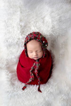 Sleeping newborn wrapped in red with festive plaid bonnet on white fur backdrop.