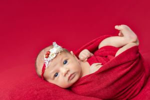 Infant wearing white headband lying on vibrant red blanket during newborn photoshoot.
