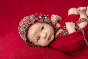 Baby wearing plaid bonnet on red background during holiday themed photoshoot.