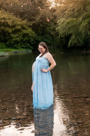 Ethereal maternity photo of an expectant mother in a flowing blue dress standing in a serene pond.