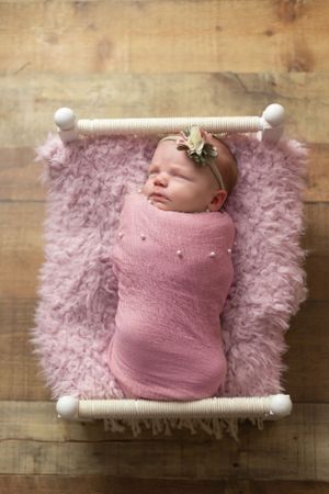 Sleeping newborn swaddled in pink wrap on fluffy pink blanket in white miniature bed.