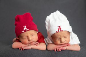 Twin newborns wearing red and white Alabama knit beanies on gray background.