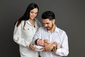 Couple in white outfits holding their newborn baby against gray background.