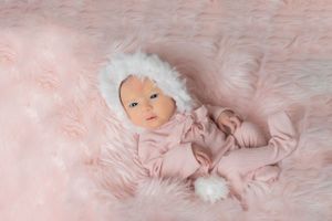 Baby in pink knit outfit lying on soft pink fur blanket with white pom pom accessories.