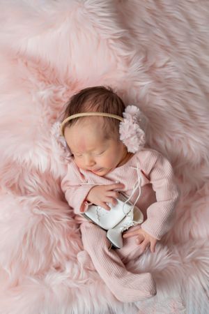 Sleeping newborn in pink outfit with flower headband holding white ice skates on pink fur.