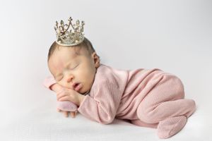 Sleeping newborn in pink outfit wearing delicate crystal crown on white background.