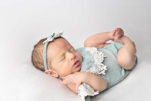 Sleeping newborn in light blue romper with lace and floral headband on white background.