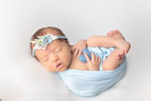 Newborn wrapped in light blue fabric with floral headband sleeping on white background.