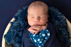 Sleeping newborn wearing blue star bib on dark background with textured fabric.