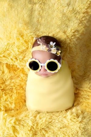 Newborn in yellow swaddle wearing sunglasses and floral headband on yellow furry backdrop.