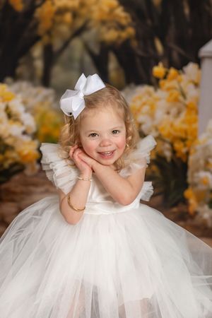 Small child wearing a white tulle dress and bow poses sweetly with hands on cheeks against autumn background.