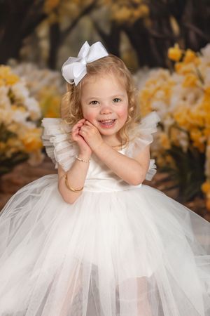 A small child in a white tulle dress and bow poses for a portrait surrounded by autumn foliage.