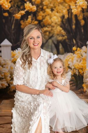Mother and daughter wearing matching white dresses pose together against autumn foliage backdrop in outdoor portrait.
