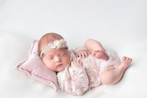 Newborn in lace outfit sleeping on pink pillow with white headband.