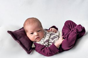 Newborn in purple outfit resting on matching pillow against white background.