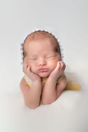 Newborn in white bonnet rests peacefully with hands under cheeks on soft white background.