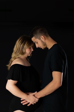 Intimate black and white portrait of an expectant couple in dark clothing against a black background.
