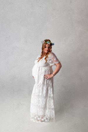 Maternity portrait featuring a white lace dress and floral crown against neutral background.