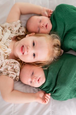 Three young siblings lying together on white bedding share a sweet moment.