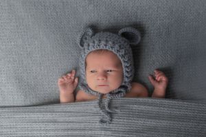 Baby wearing adorable gray knit bear hat peeks out from gray textured blanket.