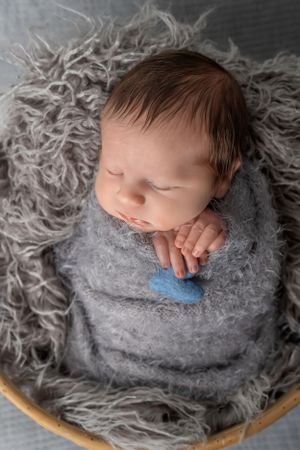 Sleeping newborn wrapped in soft gray wrap rests on textured flokati rug.