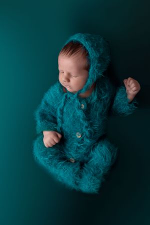 Newborn wearing teal fuzzy knit outfit sleeps peacefully against dark teal background.