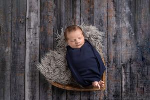 Newborn wrapped in navy swaddle rests on wooden chair against rustic wood background.