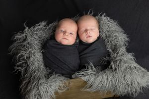 Twin newborns nestled together in black wraps on gray fur backdrop.