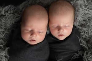Twin newborns sleeping side by side wrapped in black swaddles on gray textured backdrop.