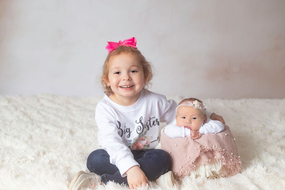 Two siblings on a white fur rug, one holding a baby doll.