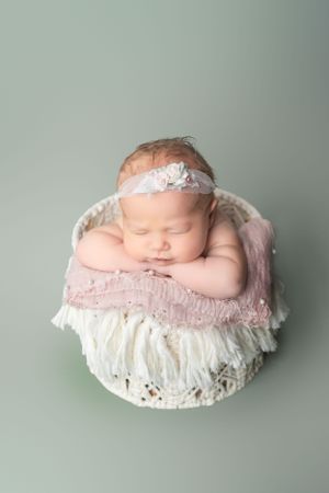 Sleeping newborn with lace headband posed in a white basket on mint green background.