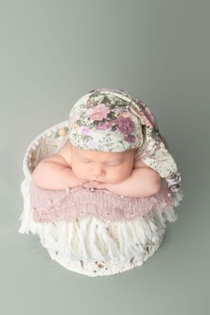 Newborn sleeping in basket wearing floral bonnet against soft green background.
