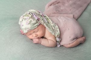 Sleeping newborn in floral bonnet and pink wrap on mint green backdrop.