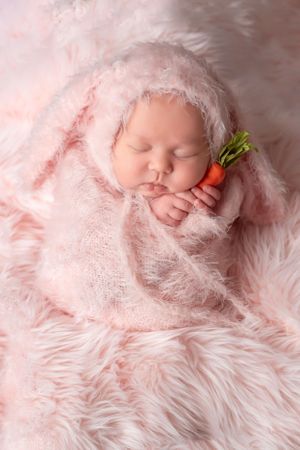 Sleeping newborn in pink fuzzy wrap with tiny decorative carrot prop.