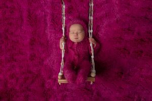 Newborn baby wrapped in burgundy fabric rests peacefully on a small wooden swing against a deep red background.