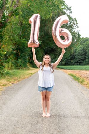 Person holding large silver number 16 balloons on a rural dirt road surrounded by trees.