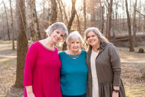 Three generations of women smile together in an outdoor autumn setting.
