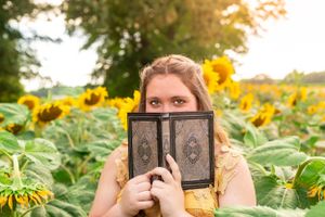 Person in yellow dress holds vintage book in front of face while standing in sunflower field.