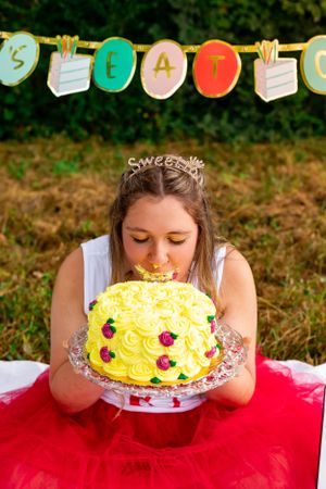 A yellow birthday cake with roses is being eaten face-first while a festive banner hangs in the background.