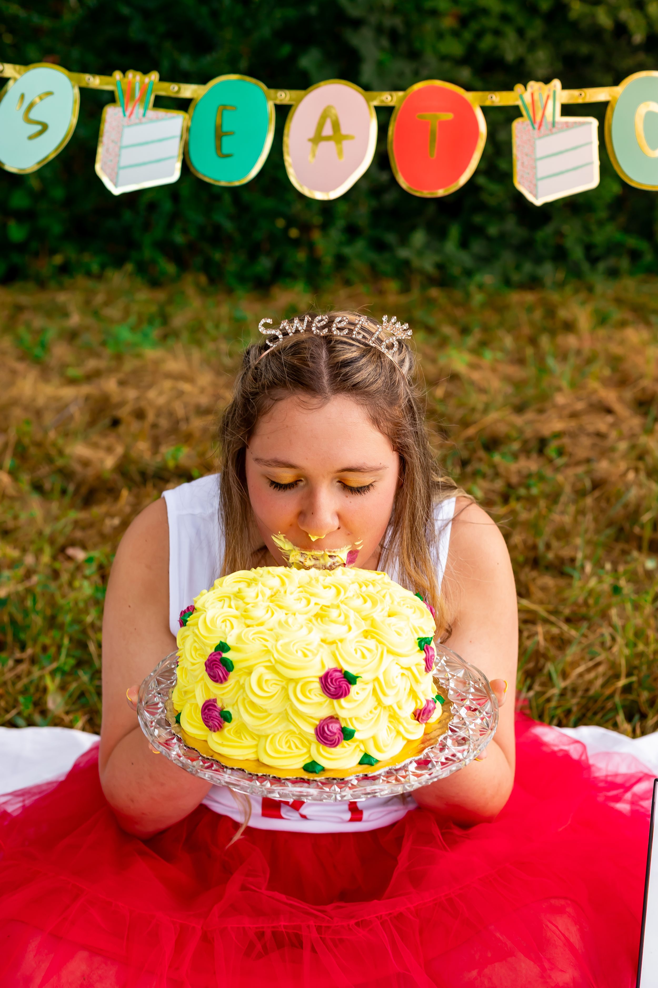 A yellow birthday cake with roses is being eaten face-first while a festive banner hangs in the background.