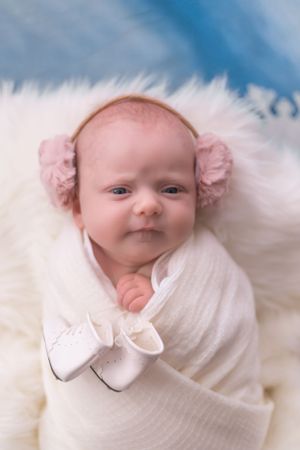 Baby wearing pink earmuffs and white wrap poses on soft white fur background.