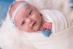 Baby in white wrap with lace headband smiles against soft blue and white background.