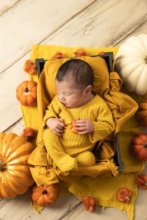 Baby in yellow knit outfit sleeps among decorative autumn pumpkins on wooden floor.