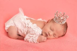 Sleeping baby wearing lace outfit and tiny crown posed on coral pink background.